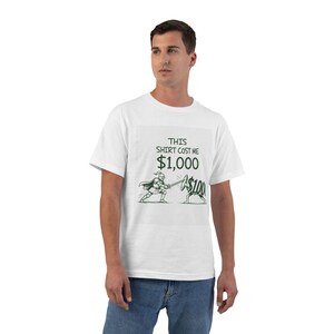 Può includere: Una t-shirt bianca a maniche corte con un'illustrazione grafica di un cavaliere che combatte contro una figura che regge uno scudo con la scritta $100. Sopra l'illustrazione, il testo recita THIS SHIRT COST ME $1,000 in lettere maiuscole verde scuro.