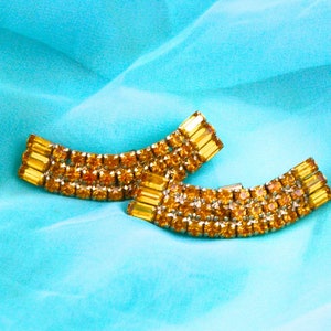 May include: A pair of gold-toned earrings with a curved design, featuring rows of sparkling amber-colored rhinestones.