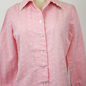 May include: A pink and white gingham button-up shirt with a white eyelet trim collar. The shirt has long sleeves and a button-up front.
