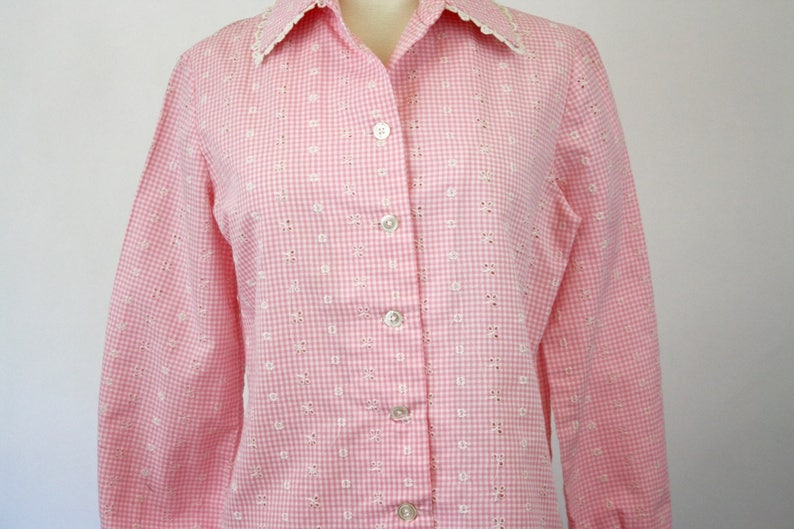 May include: A pink and white gingham button-up shirt with a white eyelet trim collar. The shirt has long sleeves and a button-up front.