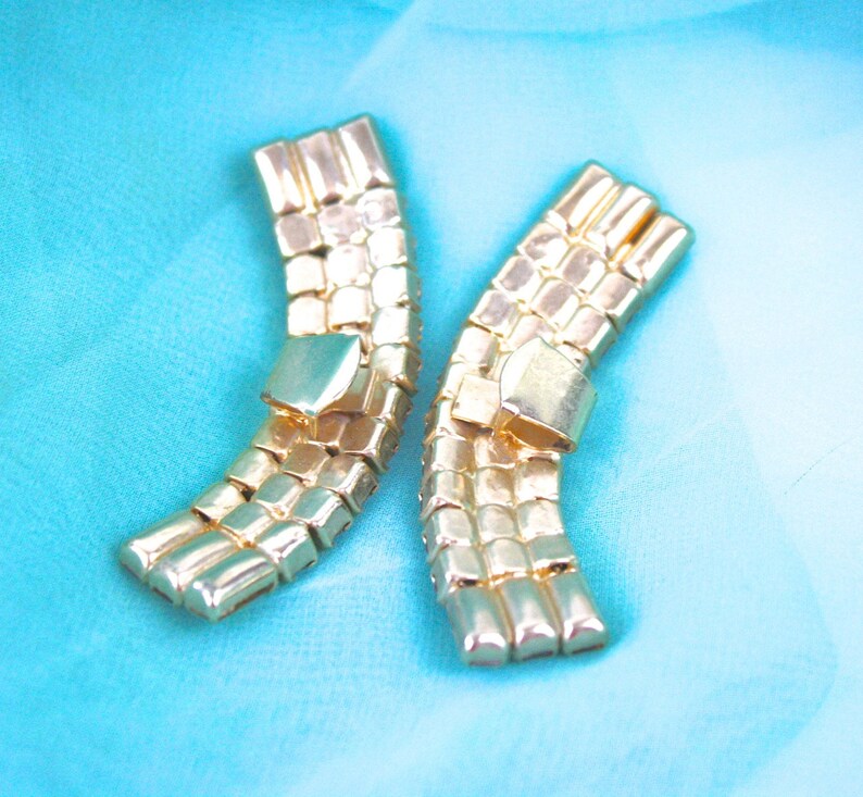 May include: Two gold-tone metal belt buckles with a geometric design. The buckles are curved and have a shiny finish.
