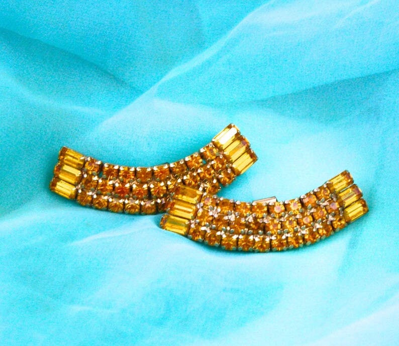 May include: A pair of gold-toned earrings with a curved design, featuring rows of sparkling amber-colored rhinestones.