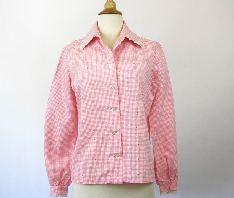 May include: A pink and white gingham button-up shirt with a white lace collar and cuffs. The shirt has a fitted silhouette and a long sleeve.