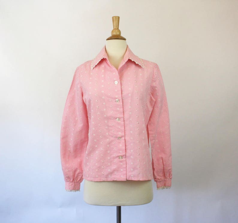 May include: A pink and white gingham button-up shirt with a white lace trim collar and cuffs. The shirt has a long sleeve and a fitted silhouette.