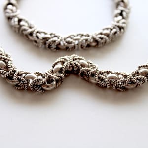 May include: A silver chain necklace with a twisted rope design. The chain is made of small, round links that are interlocked to create a rope-like effect.