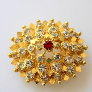 May include: A gold-toned brooch with a circular design featuring a red rhinestone in the center and surrounded by smaller clear rhinestones.