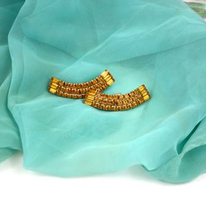 May include: A pair of gold tone earrings with amber colored rhinestones. The earrings are shaped like a curved bar and are set on a turquoise blue fabric background.