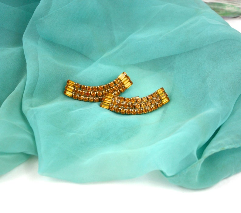 May include: A pair of gold tone earrings with amber colored rhinestones. The earrings are shaped like a curved bar and are set on a turquoise blue fabric background.