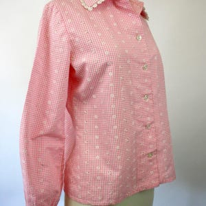 May include: A pink and white gingham button-up shirt with a white lace collar and embroidered white flowers. The shirt has long sleeves and a button-up front.