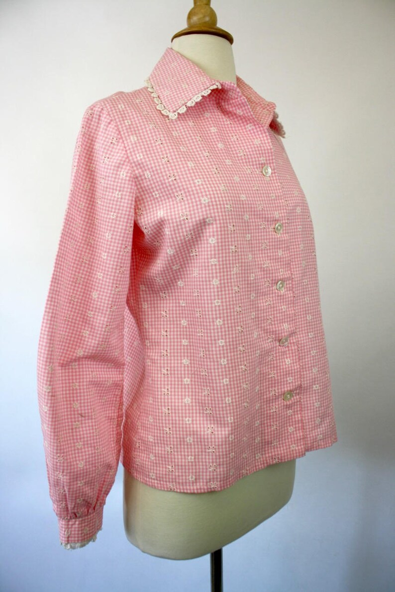 May include: A pink and white gingham button-up shirt with a white lace collar and embroidered white flowers. The shirt has long sleeves and a button-up front.
