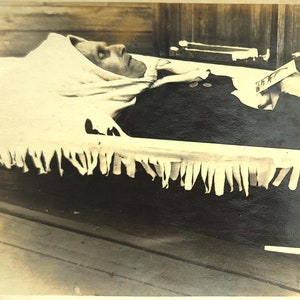 以下が含まれることがあります： 棺の中に横たわる人のヴィンテージ白黒写真。その人は白い頭飾りと暗い服を着ています。棺は装飾的なフリンジで裏打ちされています。