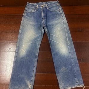 Puede incluir: Unos vaqueros de mezclilla azul con un diseño clásico de cinco bolsillos, cierre de botones y un acabado de lavado medio con desvanecimientos en los muslos y rodillas. El pantalón de corte recto presenta un ligero desgaste y bajos deshilachados.