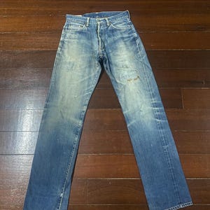 May include: Faded blue denim jeans with a button fly. The jeans show signs of wear, including fading and a small tear on the right leg. The jeans are laid flat on a wooden surface.