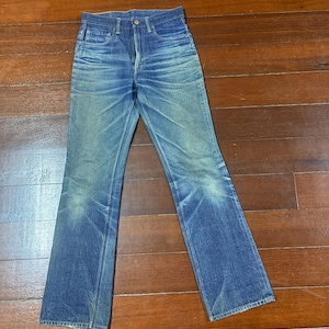 May include: Faded blue denim jeans with a classic five-pocket design. The jeans have a straight leg fit and show signs of wear and fading, giving them a vintage look. The jeans are laid flat on a wooden surface.