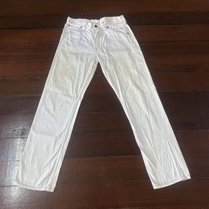 May include: A pair of white denim jeans, laid flat on a wooden surface. The jeans have a classic five-pocket design, a button and zipper closure, and a straight leg. The fabric appears slightly worn.