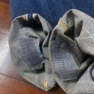 May include: Close-up of the folded hem of blue denim jeans. The cuffs are turned up, revealing the selvedge edge with red and white stripes. Patches are sewn on the inside of the cuffs. The jeans are on a wooden surface.