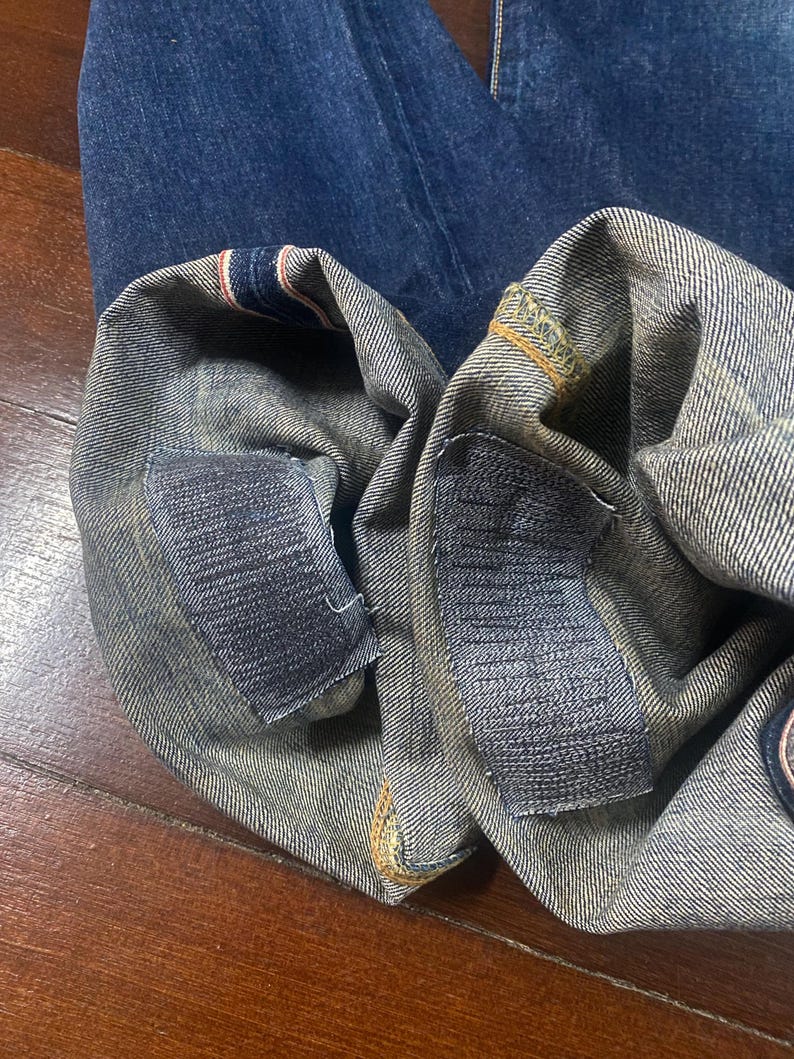 May include: Close-up of the folded hem of blue denim jeans. The cuffs are turned up, revealing the selvedge edge with red and white stripes. Patches are sewn on the inside of the cuffs. The jeans are on a wooden surface.