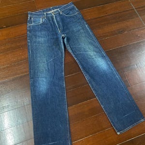 May include: A pair of blue denim jeans with a classic five-pocket design. The jeans have a button fly and a straight leg cut. The denim has a faded appearance, with lighter areas on the thighs and knees. The jeans are laid flat on a wooden surface.