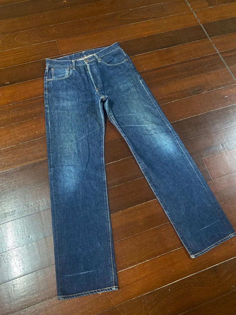 May include: A pair of blue denim jeans with a classic five-pocket design. The jeans have a button fly and a straight leg cut. The denim has a faded appearance, with lighter areas on the thighs and knees. The jeans are laid flat on a wooden surface.