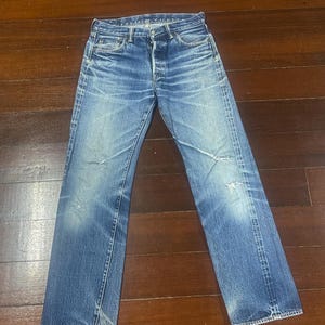 Może przedstawiać: Spodnie dżinsowe w kolorze wyblakłego niebieskiego z zapięciem na guziki i klasycznym pięciokieszeniowym krojem. Dżinsy noszą ślady zużycia z przetarciami i jasnym praniem, co nadaje im wygląd vintage. Dżinsy leżą płasko na drewnianej powierzchni.