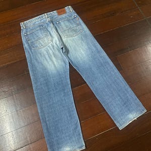 May include: Light blue denim jeans with a classic five-pocket design. The jeans have a straight leg cut and a faded wash, with a leather patch on the back waistband. The jeans are laid flat on a wooden surface.