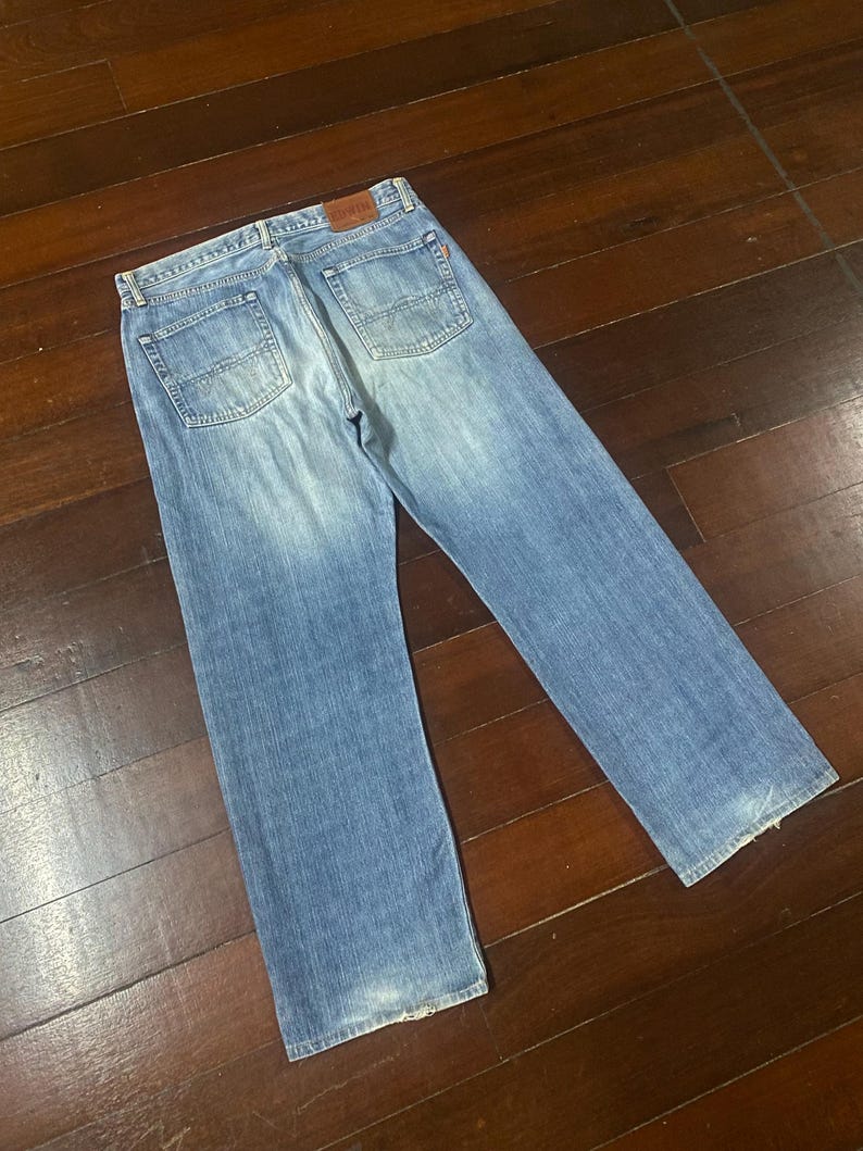 May include: Light blue denim jeans with a classic five-pocket design. The jeans have a straight leg cut and a faded wash, with a leather patch on the back waistband. The jeans are laid flat on a wooden surface.