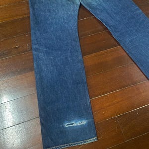 May include: Blue denim jeans with a distressed area near the hem. The jeans have a classic five-pocket design and a straight leg cut. The denim has a medium wash with subtle fading.
