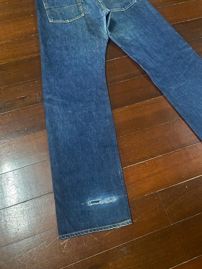May include: Blue denim jeans with a distressed area near the hem. The jeans have a classic five-pocket design and a straight leg cut. The denim has a medium wash with subtle fading.