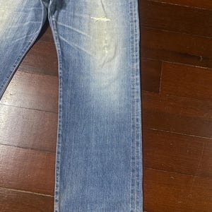 May include: Faded blue denim jeans with a distressed area on the front. The jeans have a classic five-pocket design and a straight leg cut. The denim has a worn appearance.