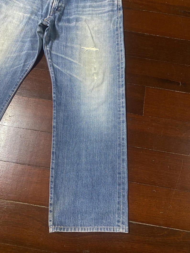 May include: Faded blue denim jeans with a distressed area on the front. The jeans have a classic five-pocket design and a straight leg cut. The denim has a worn appearance.