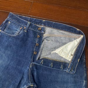 May include: Close-up of blue denim jeans with a button fly. The jeans have a classic five-pocket design and a faded wash. The button fly is partially unbuttoned, revealing the interior lining.