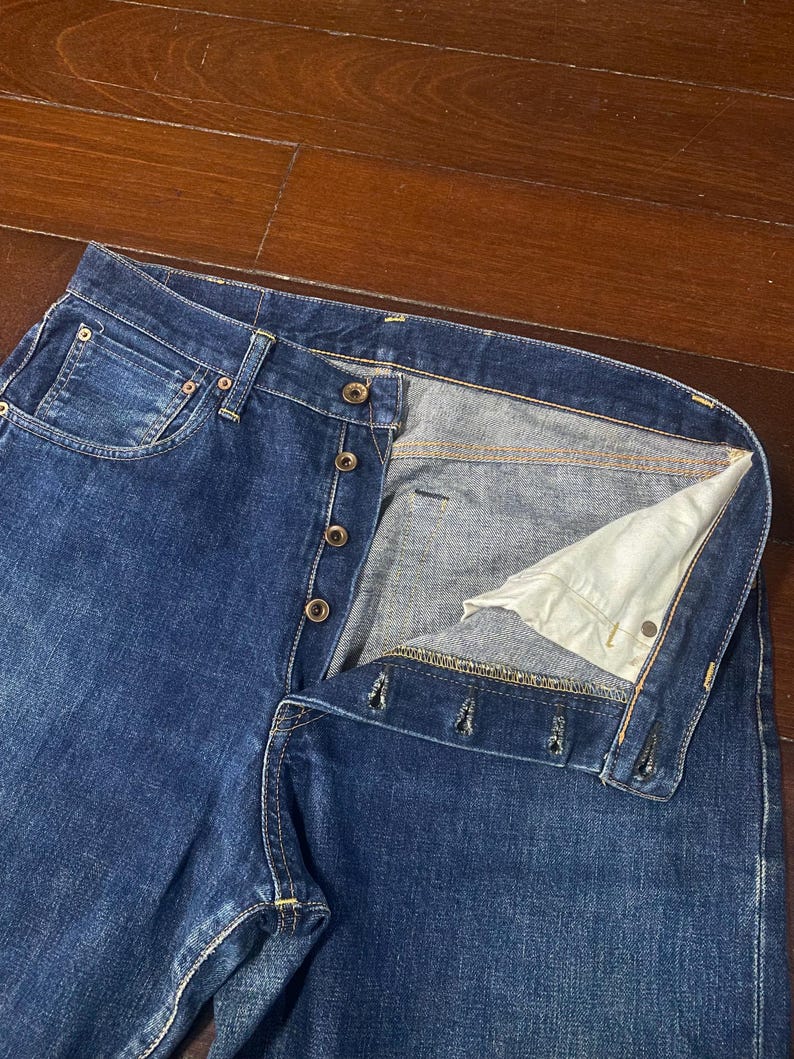 May include: Close-up of blue denim jeans with a button fly. The jeans have a classic five-pocket design and a faded wash. The button fly is partially unbuttoned, revealing the interior lining.