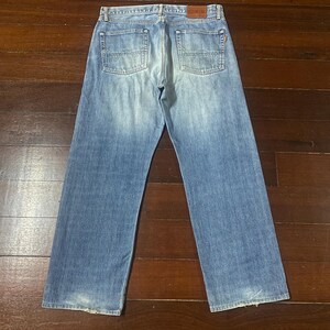 May include: Light blue denim jeans with a faded wash and a classic five-pocket design. The jeans have a straight leg and a leather patch on the back waistband. The jeans are displayed on a dark wood surface.
