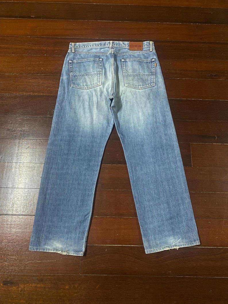May include: Light blue denim jeans with a faded wash and a classic five-pocket design. The jeans have a straight leg and a leather patch on the back waistband. The jeans are displayed on a dark wood surface.