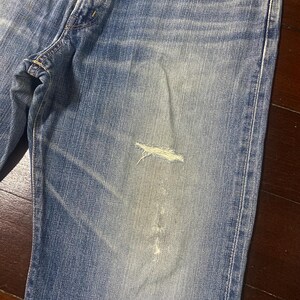 May include: Close-up of light blue denim jeans with a distressed, worn appearance. The jeans have a faded wash and a ripped section on the leg, showcasing a casual, vintage style. The fabric shows signs of wear.