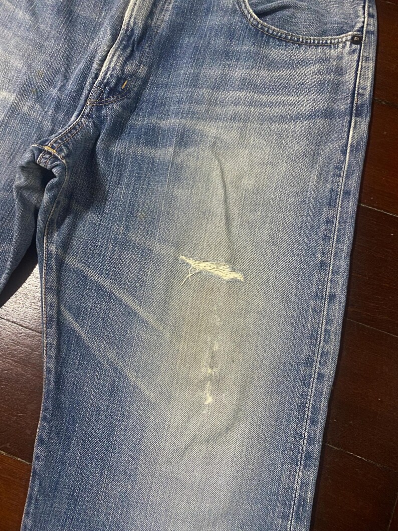 May include: Close-up of light blue denim jeans with a distressed, worn appearance. The jeans have a faded wash and a ripped section on the leg, showcasing a casual, vintage style. The fabric shows signs of wear.