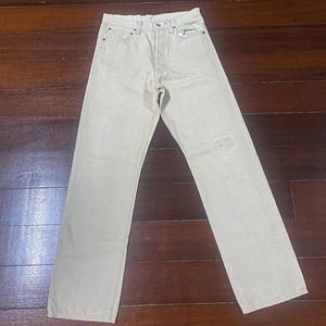 May include: Light beige denim jeans with a classic five-pocket design. The jeans have a straight leg cut and a button and zipper closure. The fabric appears to be slightly faded, giving them a vintage look. The jeans are laid flat on a wooden surface.