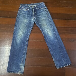 May include: Faded blue denim jeans with a classic five-pocket design. The jeans have a button fly and show signs of wear, with lighter patches on the thighs and knees. The jeans are laid flat on a wooden surface.