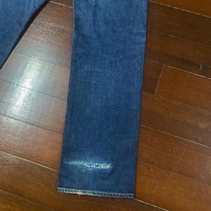 May include: Close-up of a pair of blue denim jeans. The jeans have a straight leg cut and show signs of wear, including a distressed area near the hem. The fabric has a classic, faded appearance.