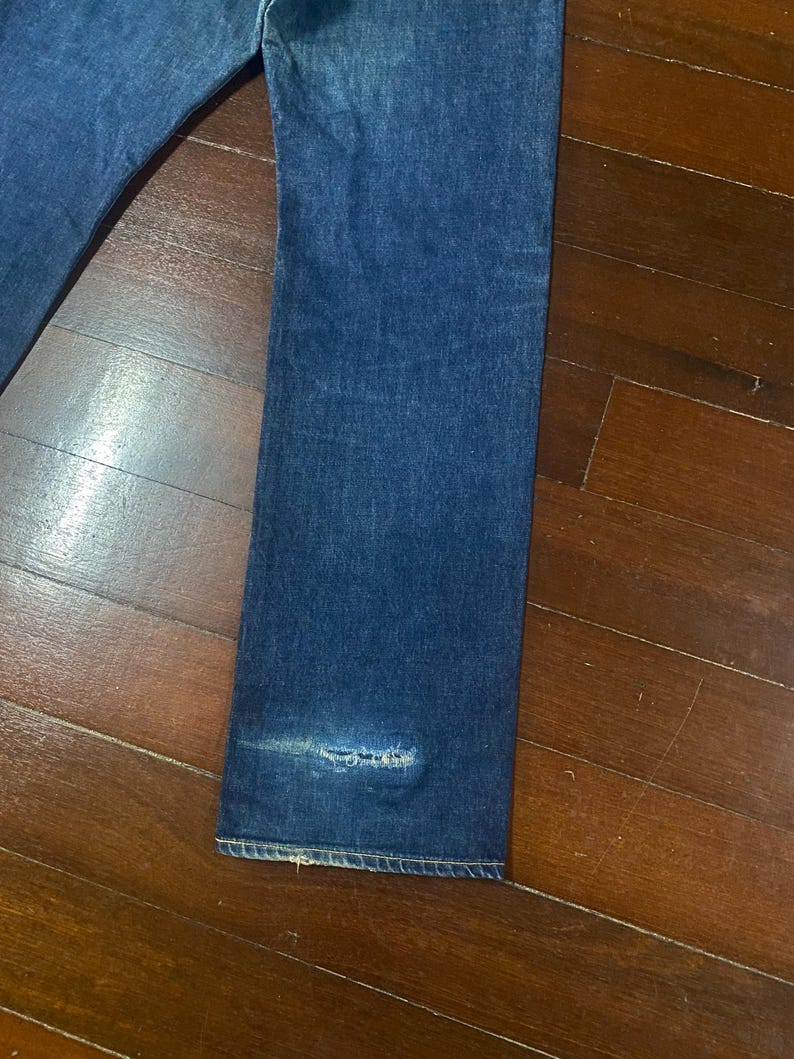 May include: Close-up of a pair of blue denim jeans. The jeans have a straight leg cut and show signs of wear, including a distressed area near the hem. The fabric has a classic, faded appearance.