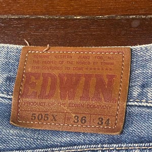 May include: A brown leather label on blue denim jeans. The label displays the brand name "EDWIN" in red. The jeans have a 36-inch waist (approx. 91 cm) and a 34-inch inseam (approx. 86 cm).