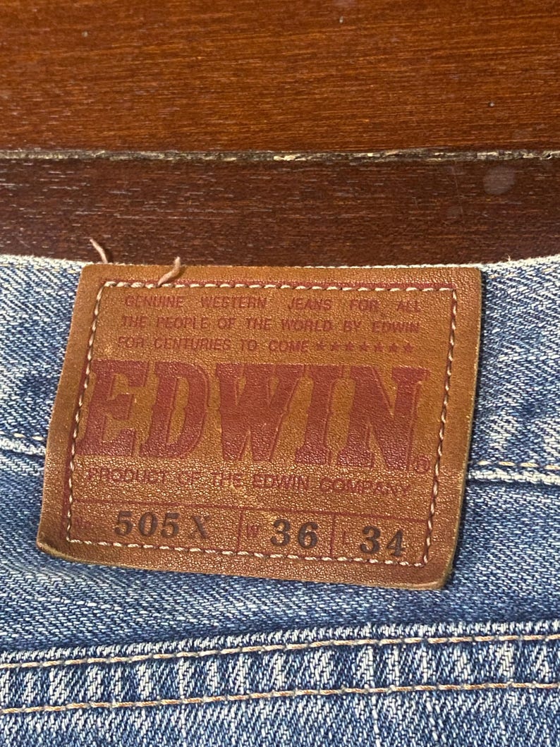 May include: A brown leather label on blue denim jeans. The label displays the brand name "EDWIN" in red. The jeans have a 36-inch waist (approx. 91 cm) and a 34-inch inseam (approx. 86 cm).