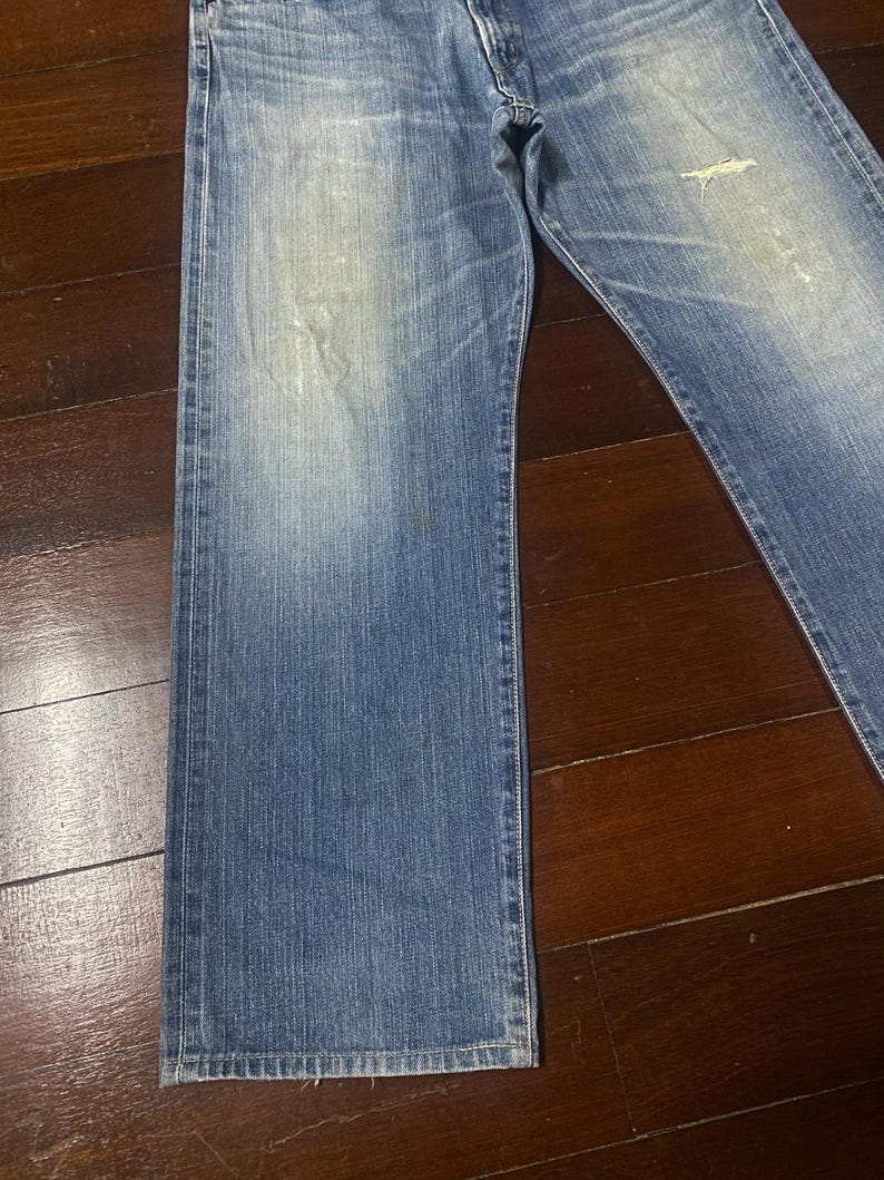 May include: Faded blue denim jeans with a classic straight leg cut. The jeans show signs of wear with distressed areas and light fading throughout. The denim has a medium weight and a traditional five-pocket design.