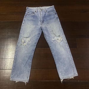 May include: Light blue denim jeans with distressed details, including ripped knees and frayed hems. The jeans have a classic five-pocket design and a button and zipper closure. The jeans are laid flat on a dark wood surface.