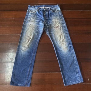 May include: Faded blue denim jeans with button fly closure. The jeans show signs of wear with lighter patches and subtle distressing. The classic five-pocket design is visible. The jeans are laid flat on a wooden surface.