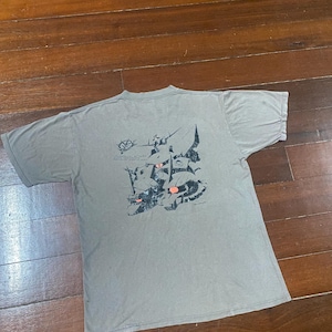 May include: A gray t-shirt with a graphic design on the back. The design features black and red illustrations of figures and text. The shirt is laid flat on a wooden surface.