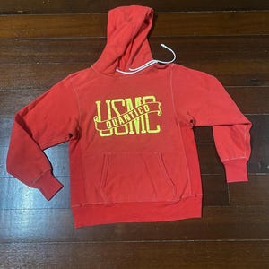 May include: Red hoodie with a kangaroo pocket and drawstring hood. The front features the text "USMC QUANTICO" in yellow lettering. The hoodie has ribbed cuffs and a ribbed hem. The garment is made of a soft, worn material.