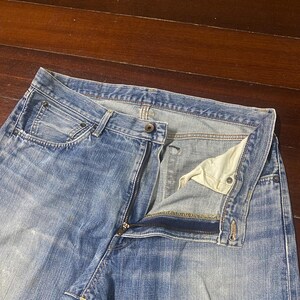 May include: Light blue denim jeans with a partially unzipped fly. The jeans have a classic five-pocket design and a faded wash. The zipper is silver-toned, and the interior lining is a light beige colour. The jeans are laid flat on a wooden surface.