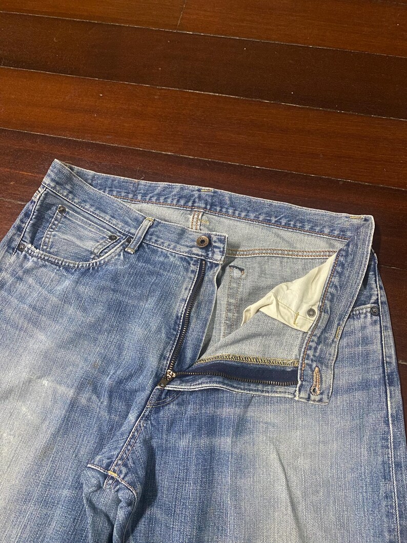 May include: Light blue denim jeans with a partially unzipped fly. The jeans have a classic five-pocket design and a faded wash. The zipper is silver-toned, and the interior lining is a light beige colour. The jeans are laid flat on a wooden surface.