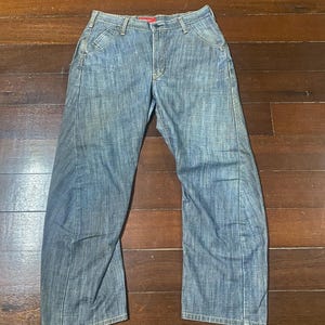 May include: Blue denim jeans with a classic five-pocket design. The jeans have a straight leg cut and a faded wash, giving them a worn-in look. The waistband features a red tag. The jeans are laid flat on a wooden surface.